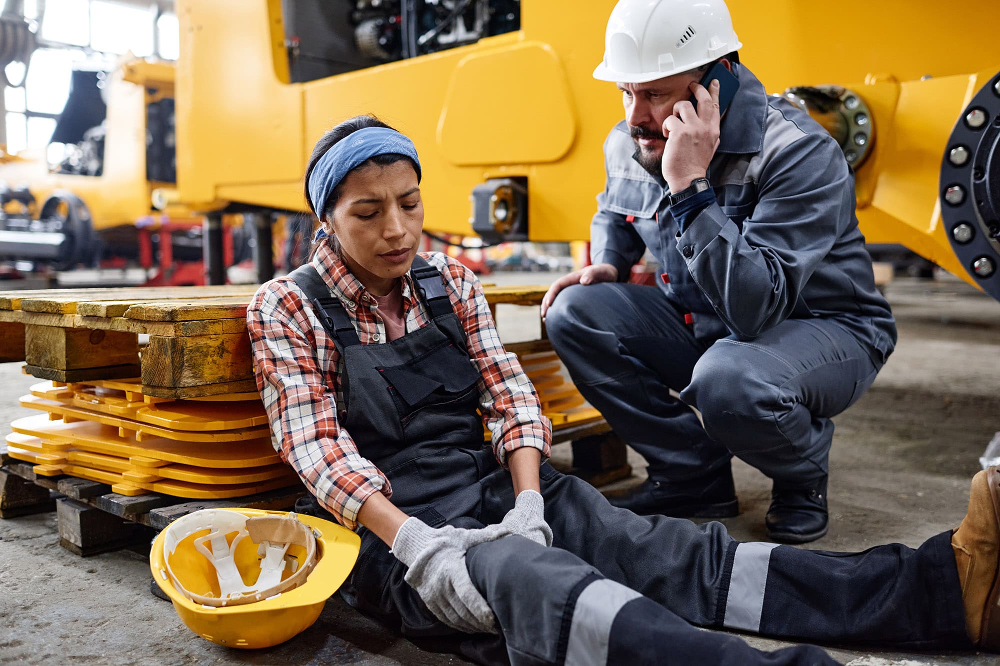 Young female engineer with hurting knee sitting by man calling ambulance