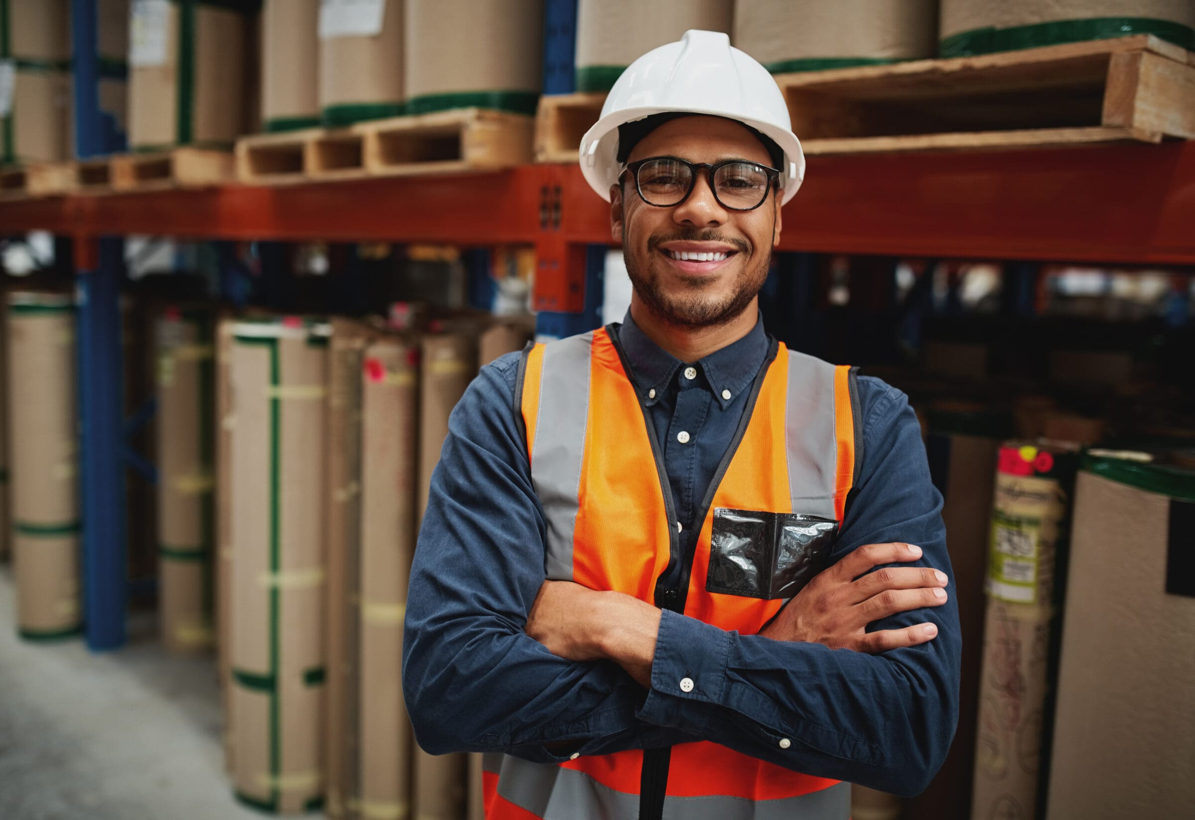 Portrait smiling worker wearing orange vest and white helmet with arms crossed in a large warehouse smiling Happy manager in warehouse standing looking at camera with folded hands